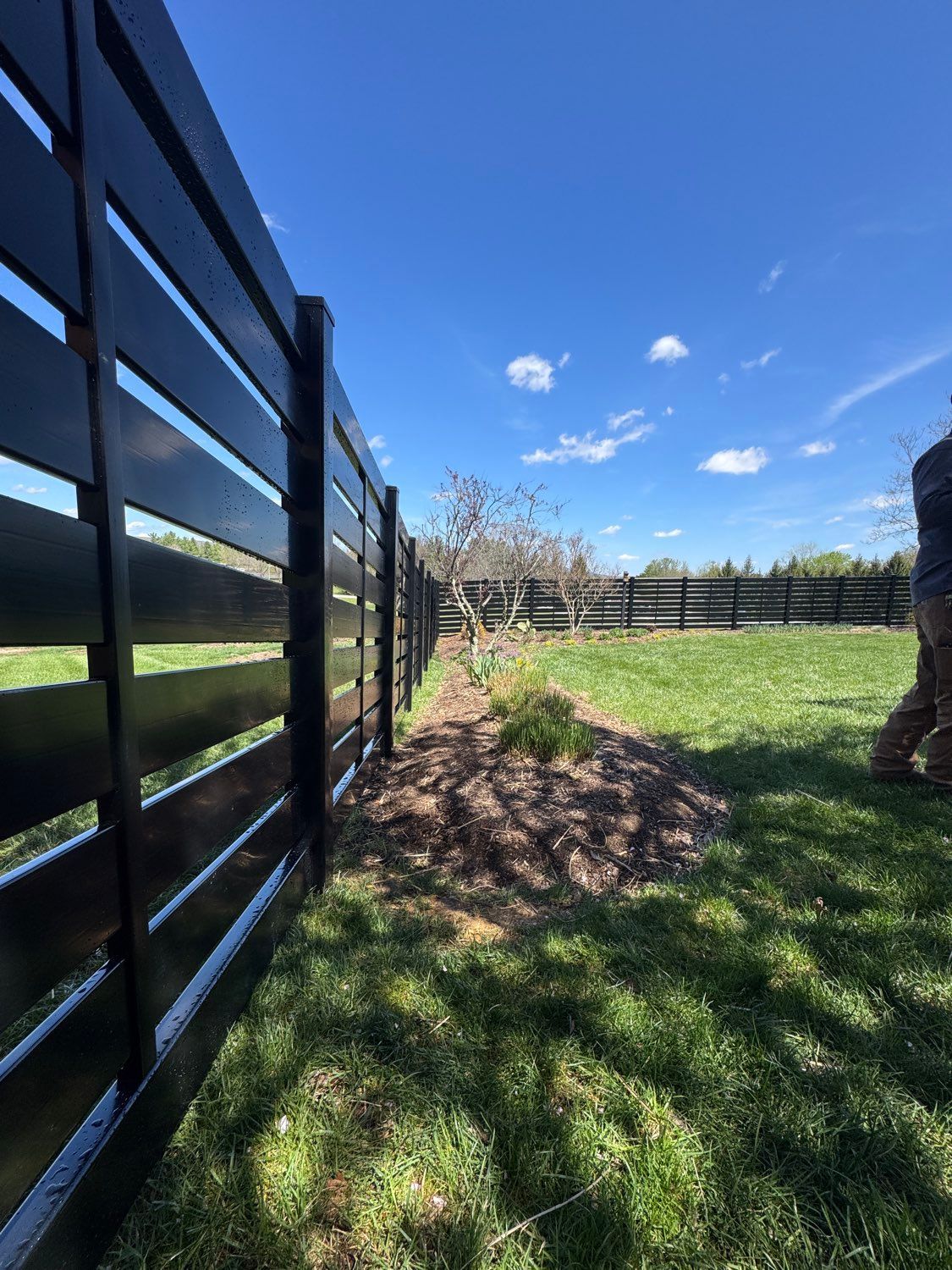 Black wooden fence beside a grassy yard under a bright blue sky with a few trees and shrubs.