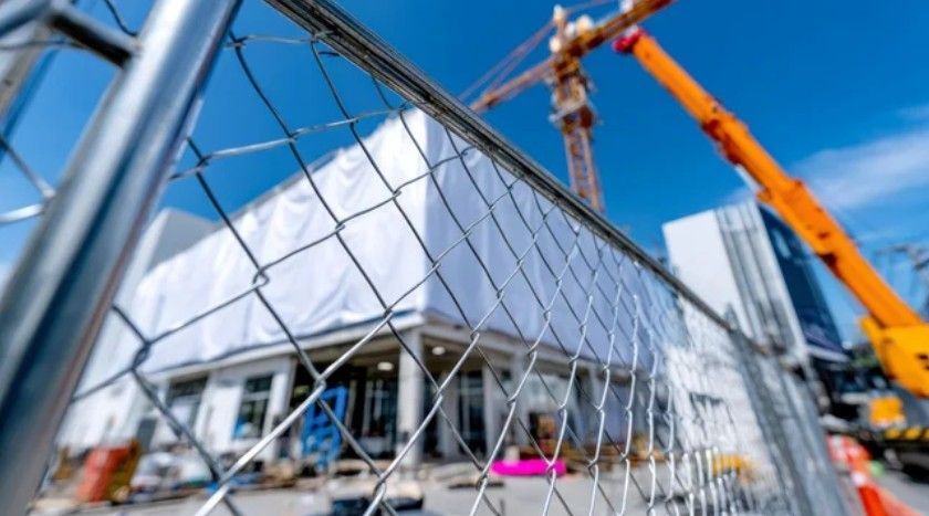 Construction site seen through a chain-link fence, with a crane and partially built building under blue sky.