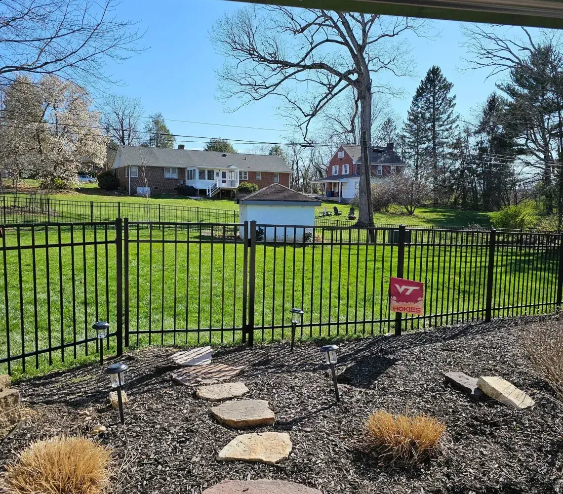 Backyard view through black fence, with stone path, green lawn, trees, and distant houses under blue sky