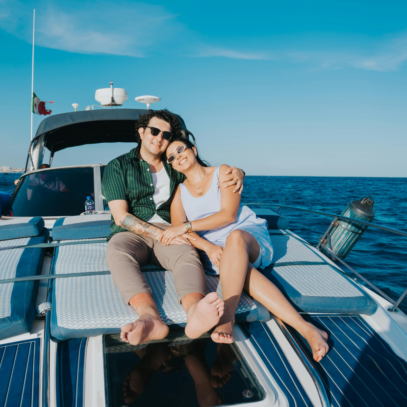 Couple sitting on the bow of a yacht, smiling on a sunny day at sea.