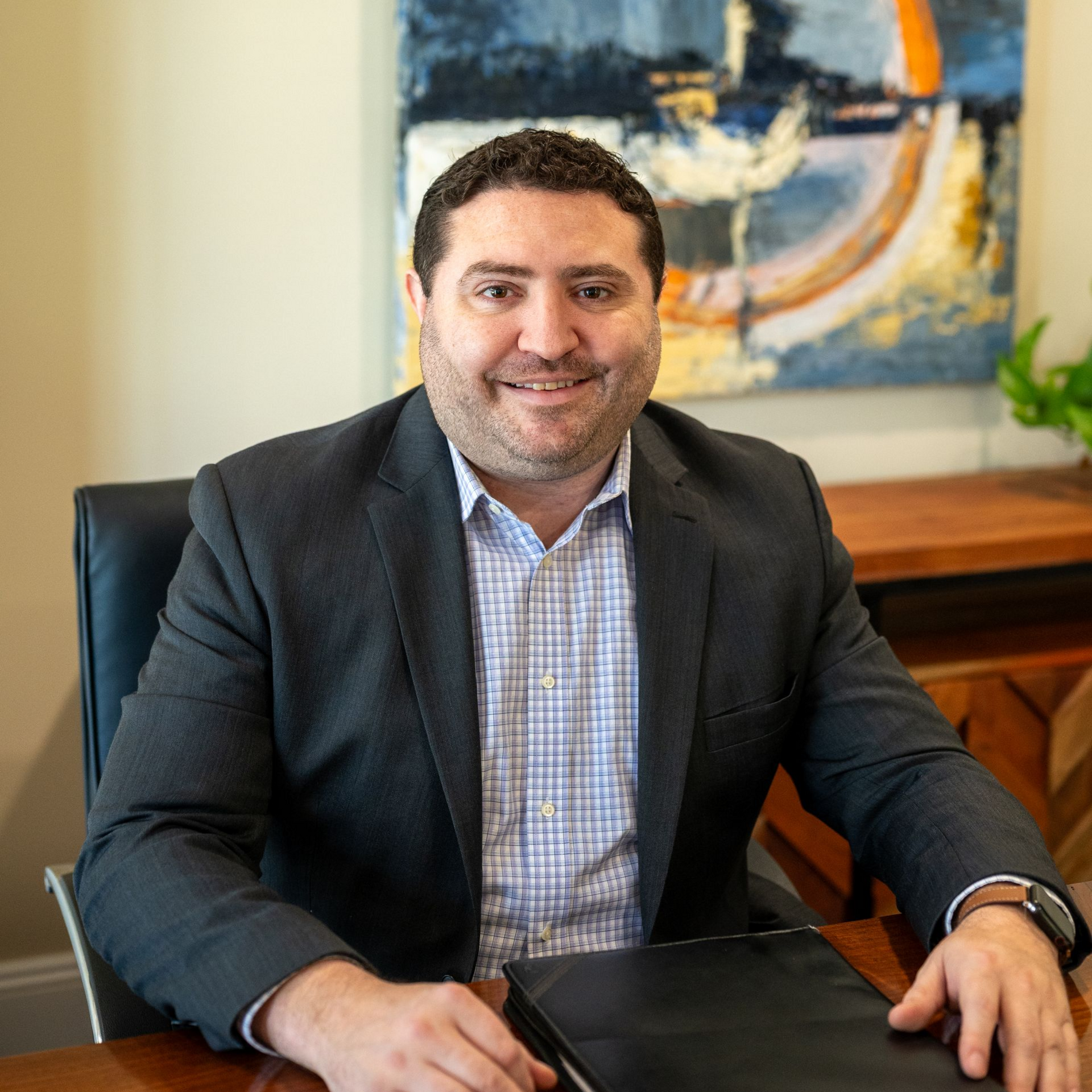 Brent Bonstingl of Delaware Wealth Planners in a dark blazer seated at a desk in an office, with an abstract painting behind him.