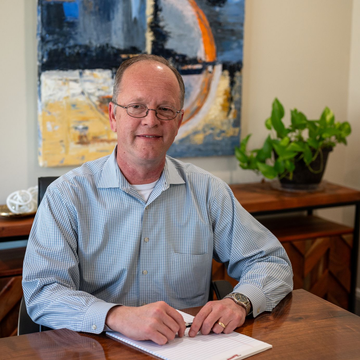 Damon Nickle of Delaware Wealth Planners seated at a wooden table in a bright room, with abstract art and a potted plant in the background