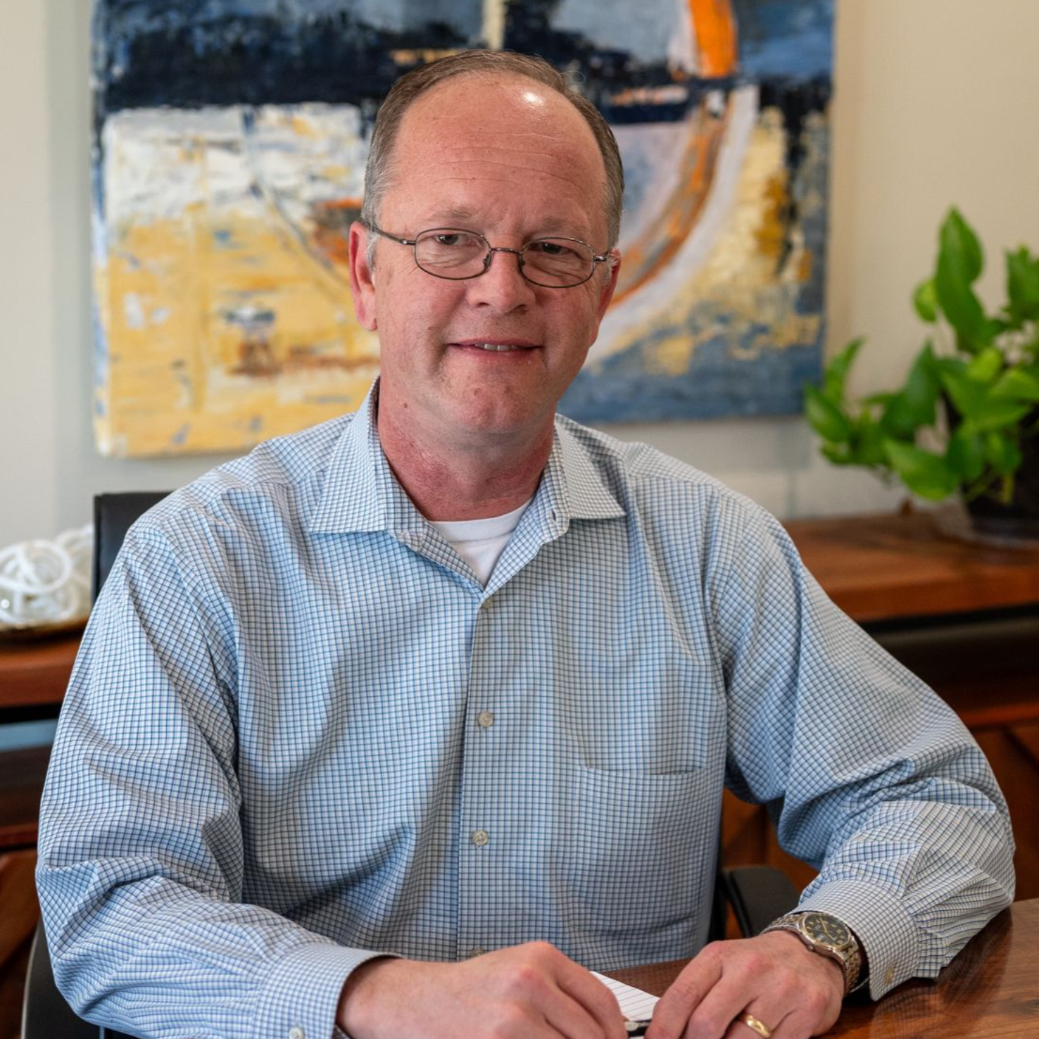 Damon Nickle of Delaware Wealth Planners seated at a desk in a light blue shirt, smiling in an office with abstract art and a plant behind him.