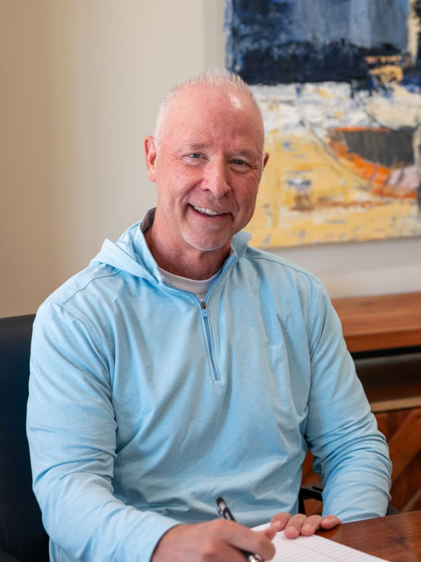 Tim Humphreys of Delaware Wealth Planners in a light blue shirt sitting at a table with a pen, in front of an abstract painting