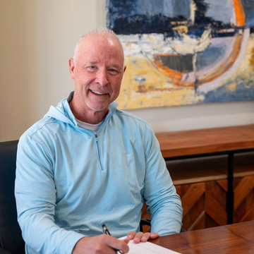 Tim Humphreys of Delaware Wealth Planners in a light blue shirt seated at a table with a pen, in front of abstract wall art.