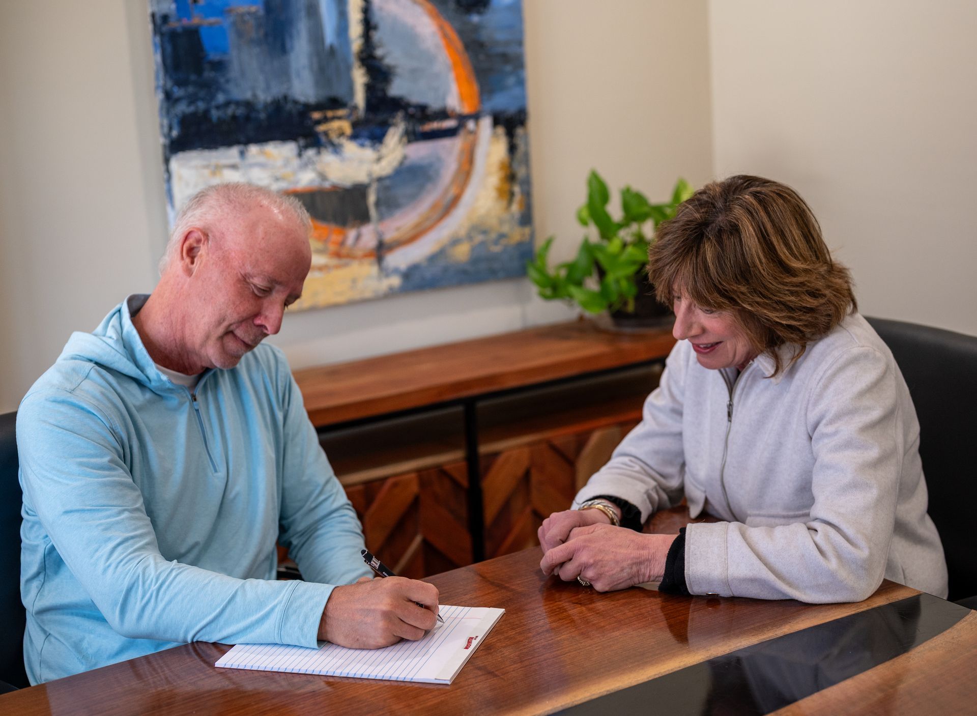 Two people sitting at a table, one writing on paper while the other watches in a bright room.