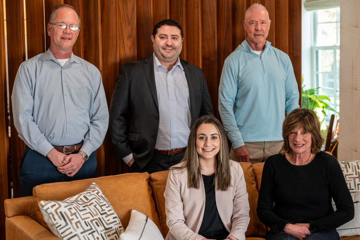 The Delaware Wealth Planners team of Tim Humphreys, Brent Bonstigl, Corrine Curcio, and Jane Atkins. Five people posing in a living room, with two seated on a couch and three standing behind them.