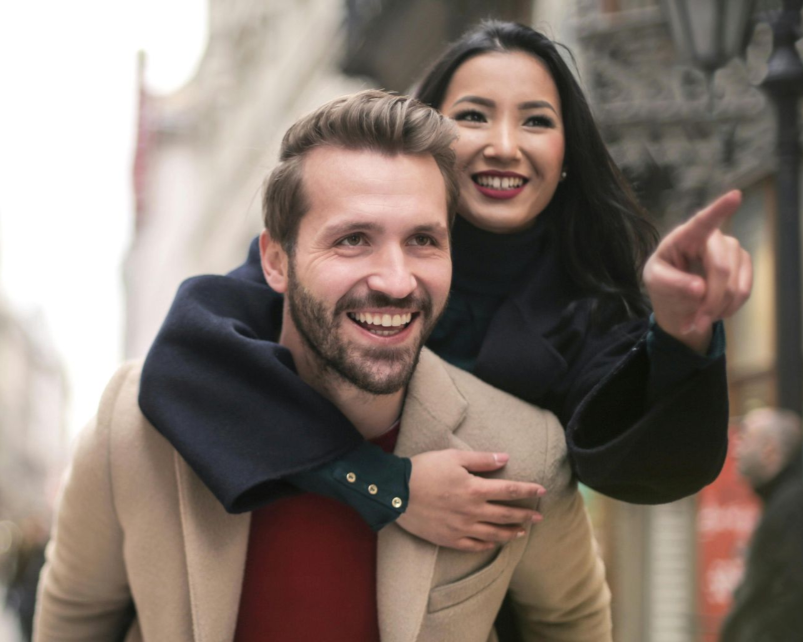 Smiling couple outdoors, the woman riding on the man’s back and pointing ahead.