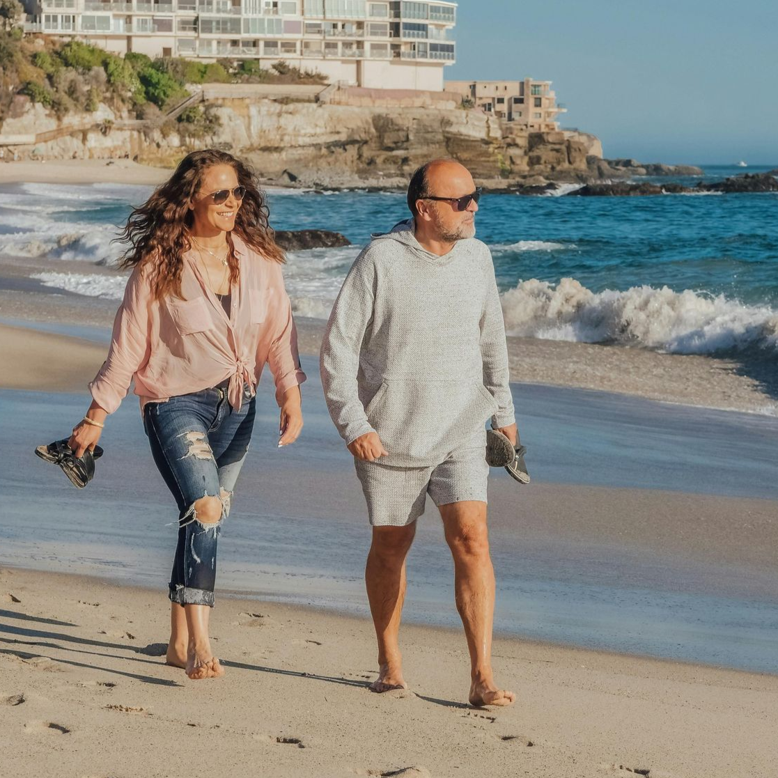 Two people walking barefoot along a sunny beach near the shoreline, with waves and seaside buildings behind them.