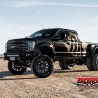 Black lifted pickup truck with oversized off-road tires parked on a dirt lot under a blue sky