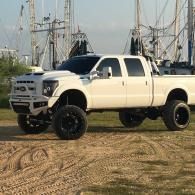 White lifted pickup truck on dirt in a marina with sailboat masts in the background