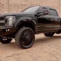 Black lifted pickup truck with large off-road tires parked on cracked pavement by a brick wall