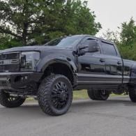 Black lifted pickup truck with oversized wheels parked on pavement near trees