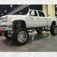 White lifted pickup truck with oversized chrome wheels displayed indoors at an auto show