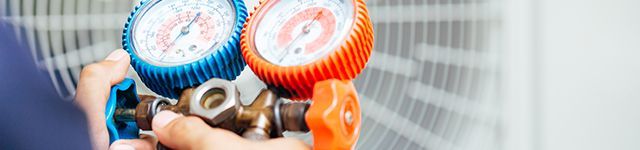 Hands holding a blue and orange pressure gauge on a metal valve indoors