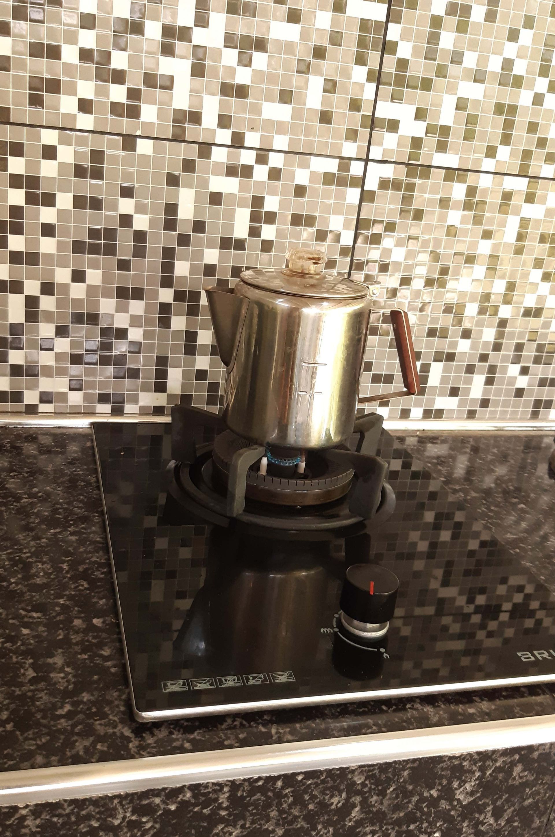 Stainless steel coffee pot on a gas stove, in front of a mosaic tile backsplash.
