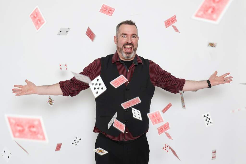 A man is standing in front of a wall with playing cards falling around him.