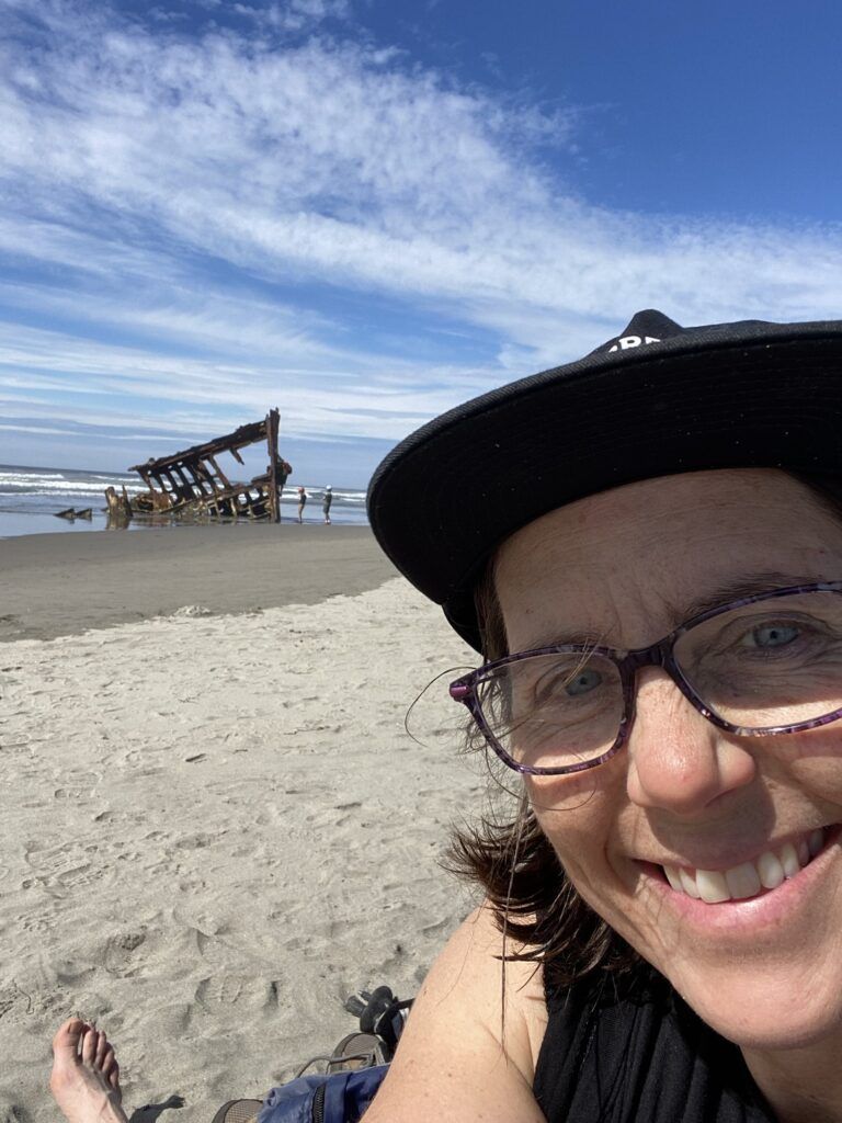A woman wearing glasses and a hat is smiling on the beach.