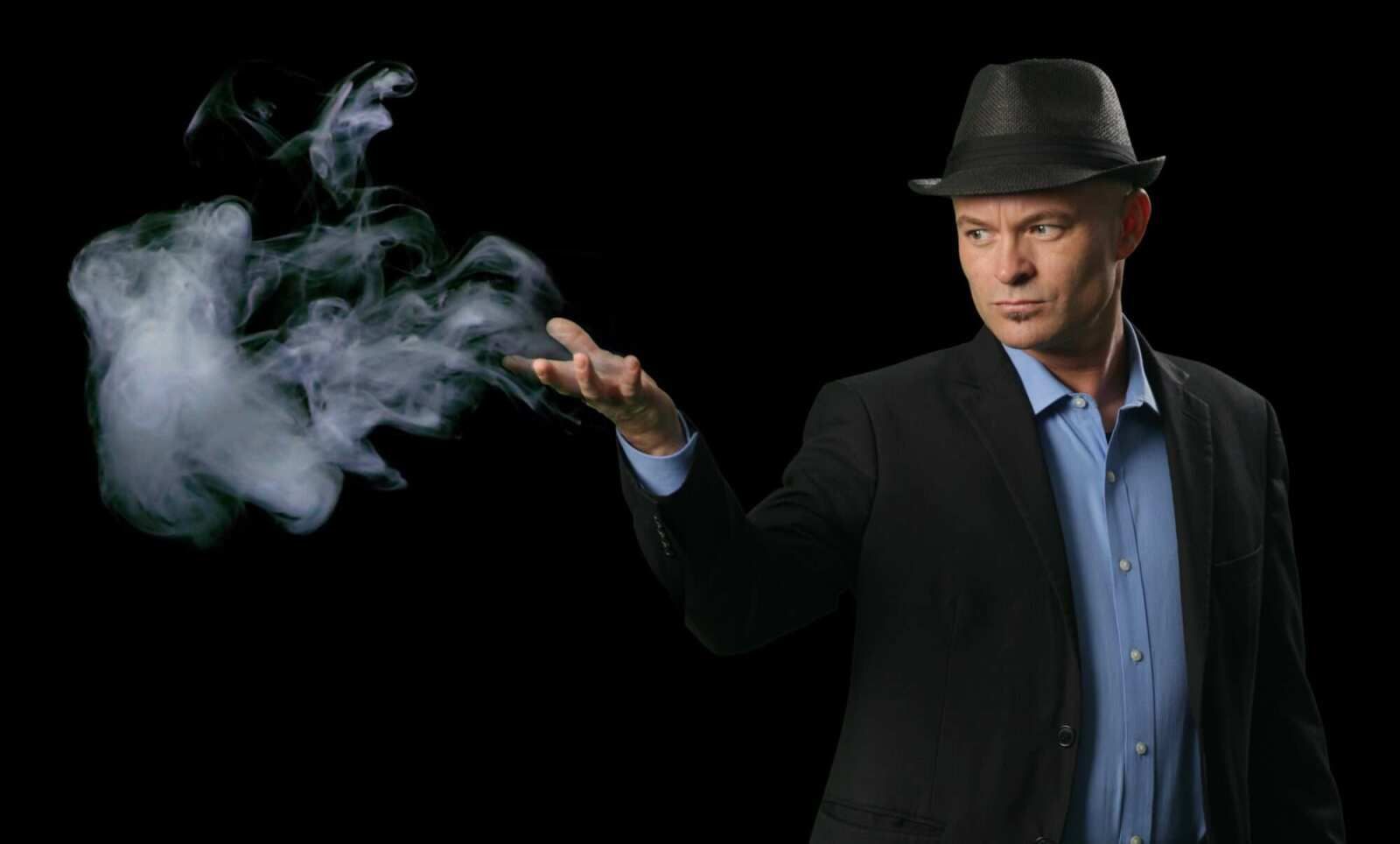 A man in a suit and hat is blowing smoke out of his hand.