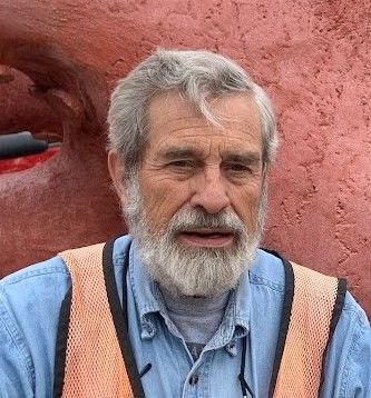 A man with a beard and an orange vest is standing in front of a red wall.