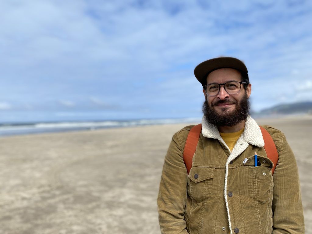 A man with a beard and glasses is standing on a beach.