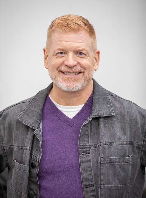 A man wearing a purple sweater and a denim jacket is smiling for the camera.