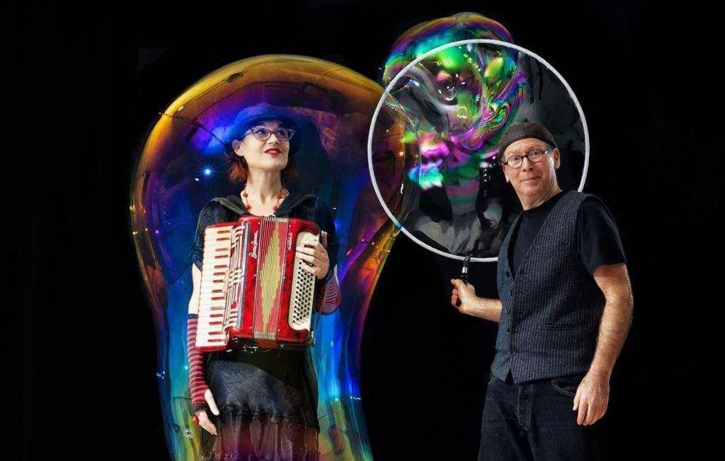 A man and a woman are playing an accordion and blowing soap bubbles.