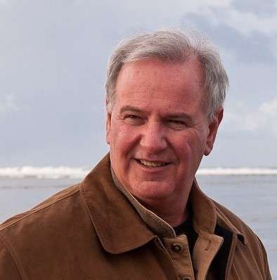 A man in a brown jacket is smiling in front of the ocean.