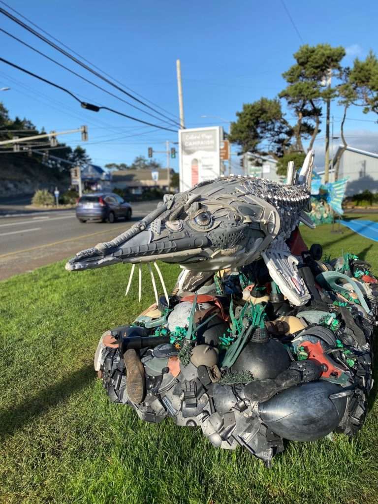 A statue of a whale made out of trash is sitting on the side of the road.