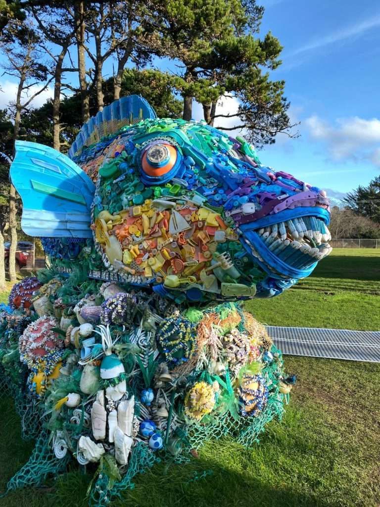 A statue of a fish made out of plastic waste in a park.