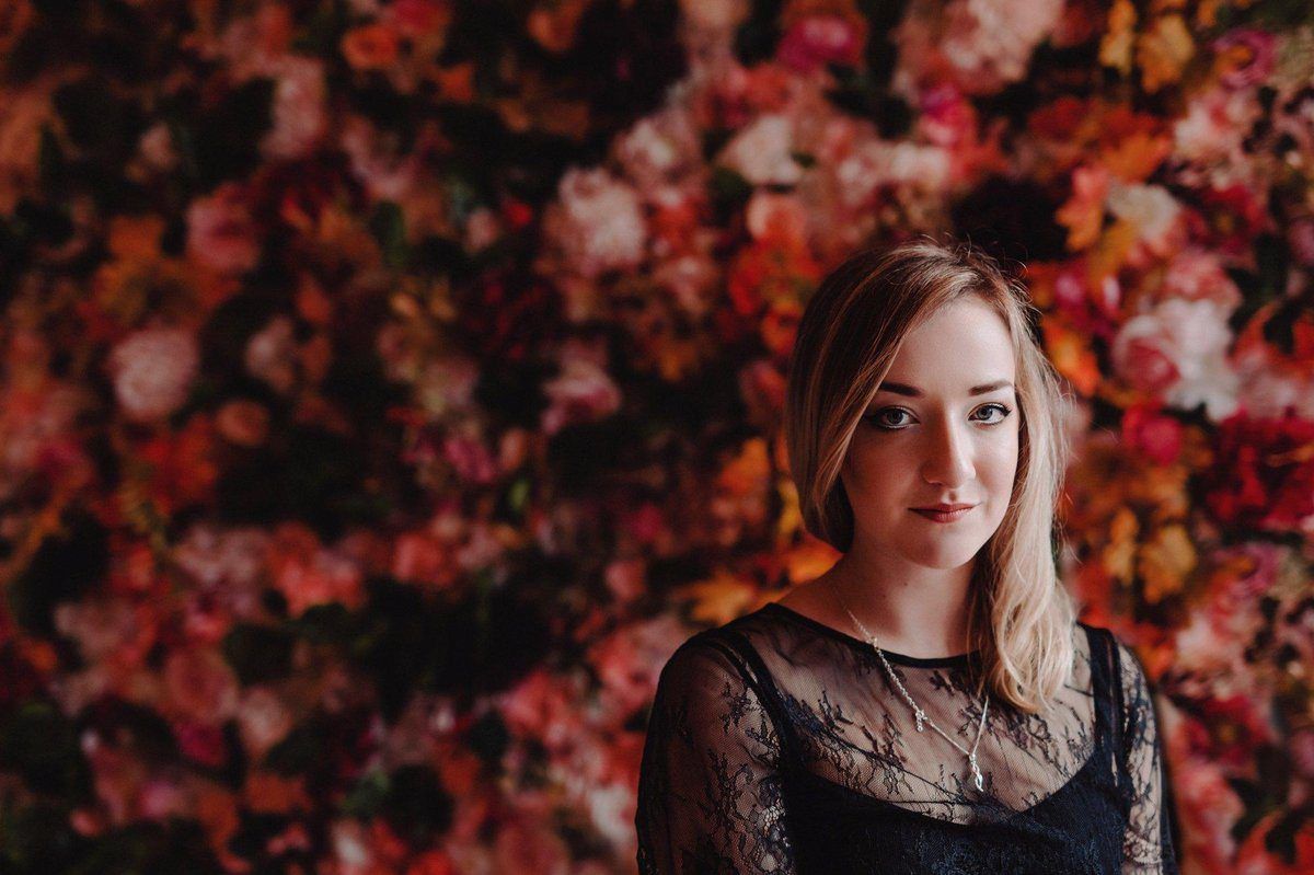 A woman in a black dress is standing in front of a wall of flowers.