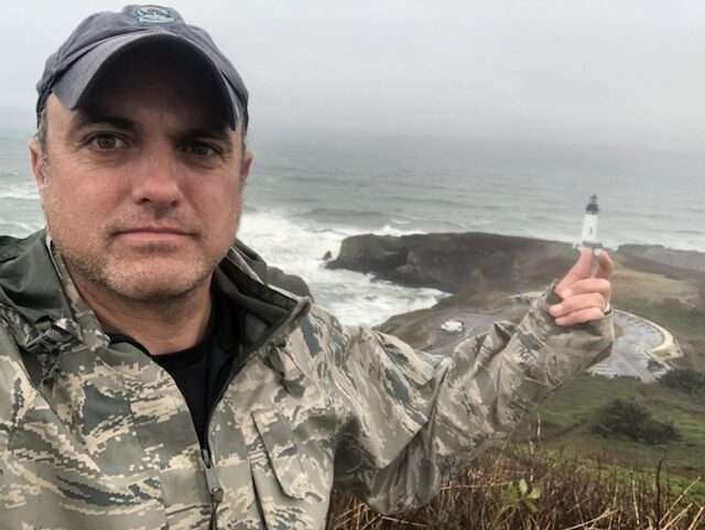 A man in a camouflage jacket is holding a lighthouse in front of the ocean.