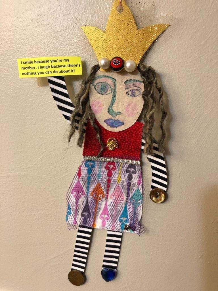 A paper doll with a crown on her head is hanging on a wall.