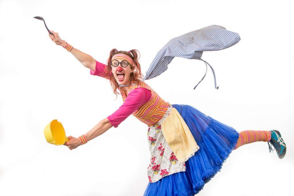 A woman in a clown costume is flying through the air holding a ladle and a pot.