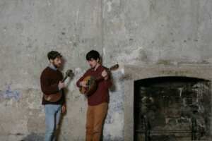 Two men are playing guitars in front of a fireplace.