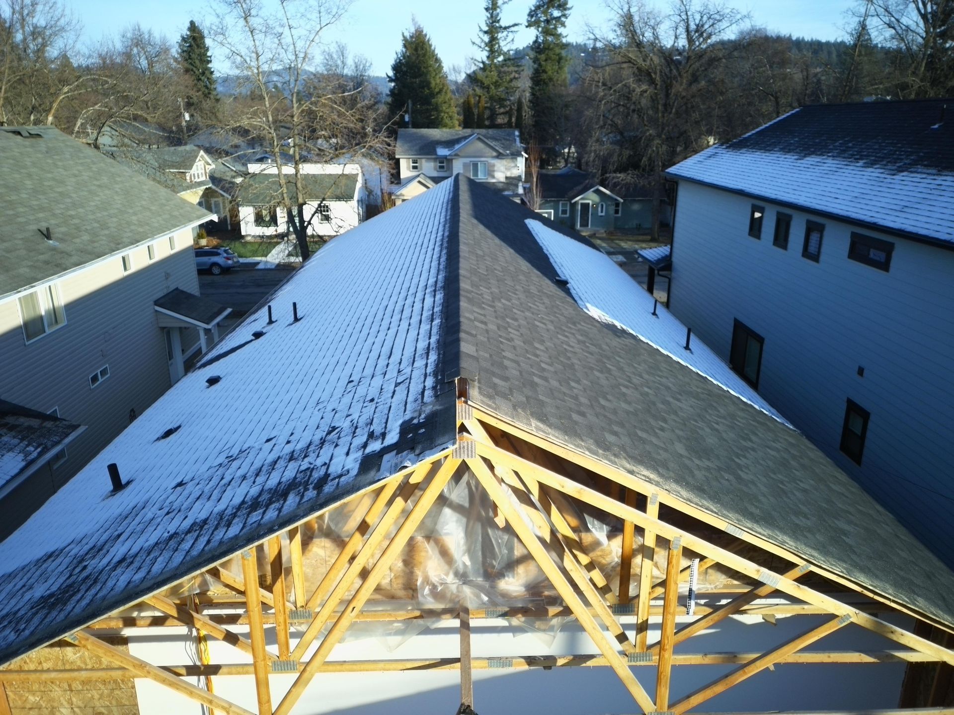 View of a house roof with snow, showing wooden trusses and surrounding houses under a bright sky.