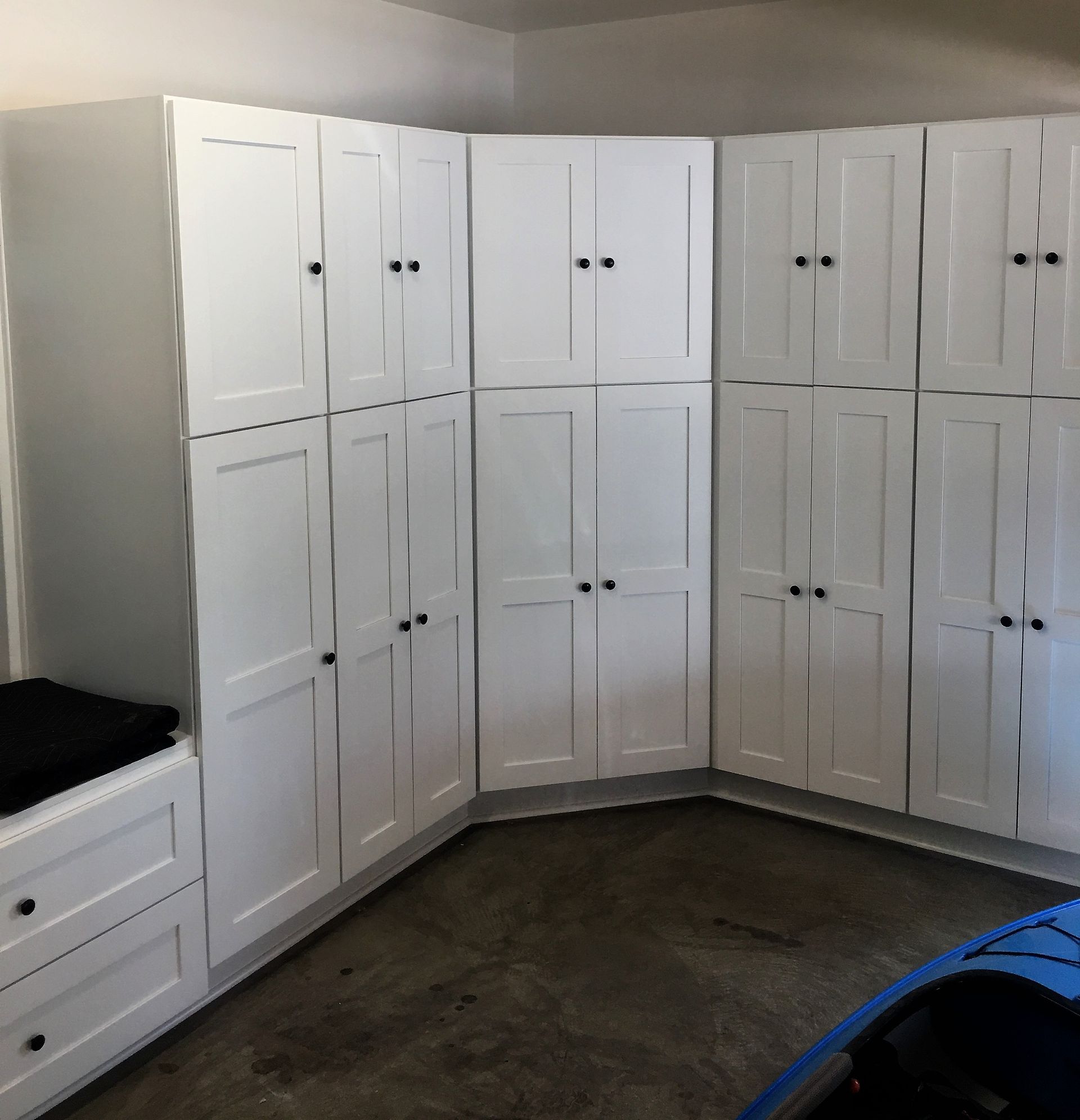 White storage cabinets in a corner of a garage, with black knobs and a blue kayak.
