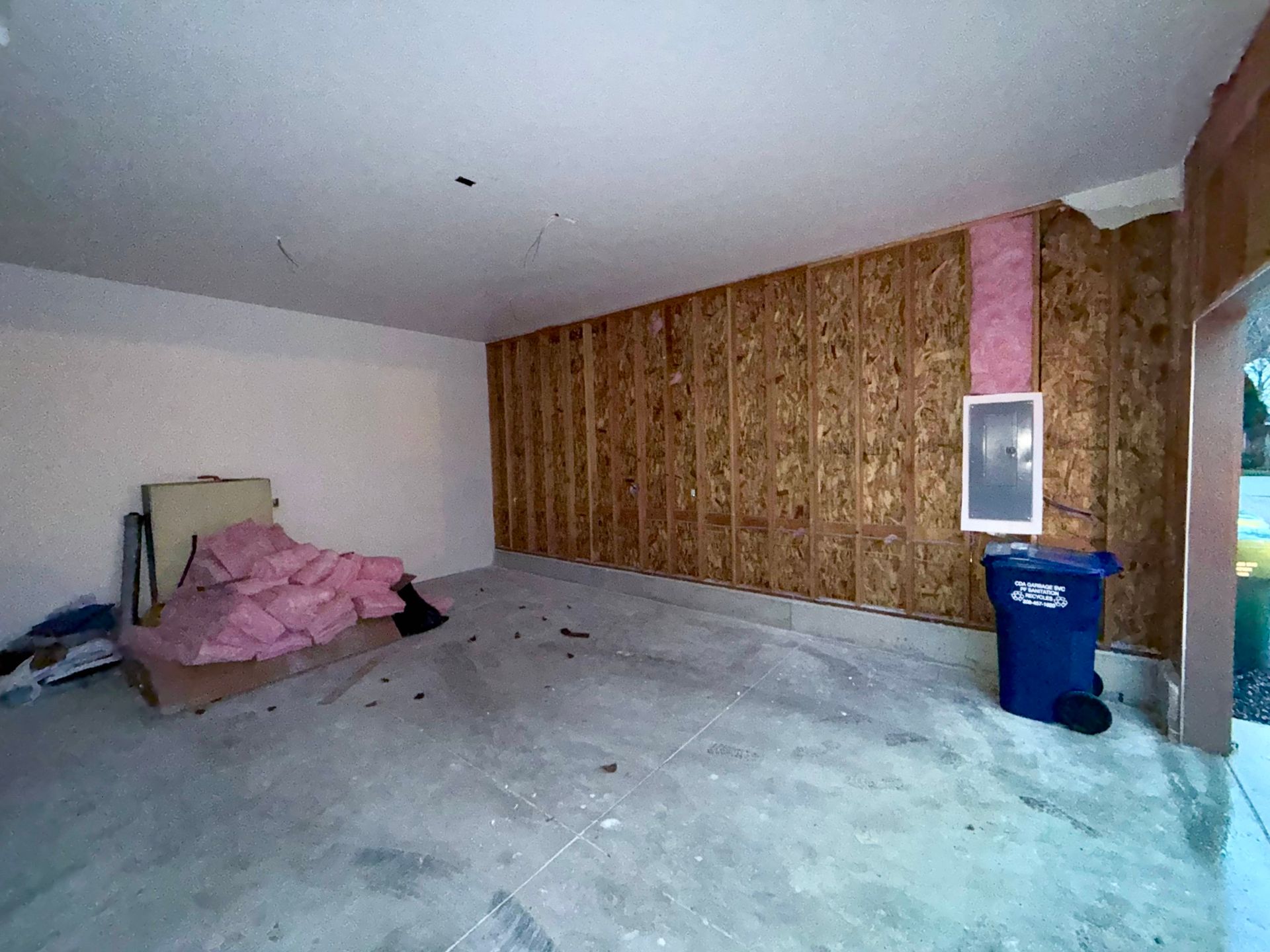 Empty garage with exposed wall studs, insulation, electrical panel, and a blue trash can.