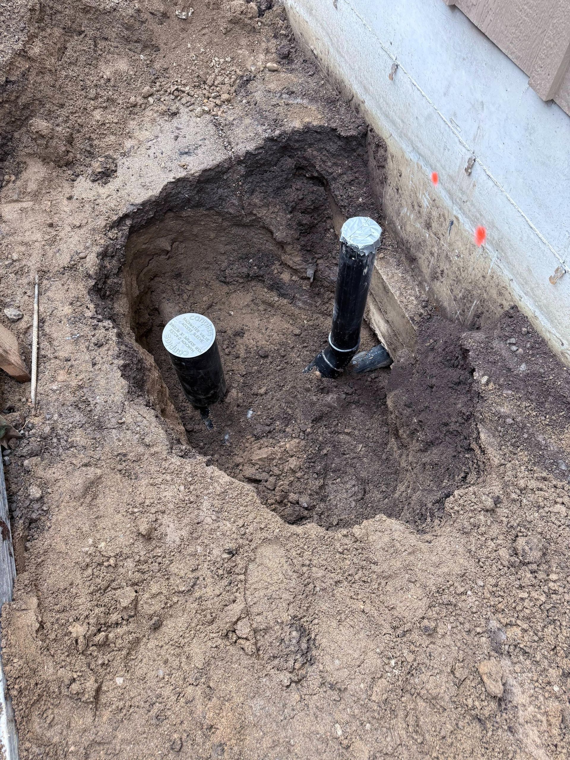 Hole dug in soil containing two black pipes with silver tops near a concrete edge.