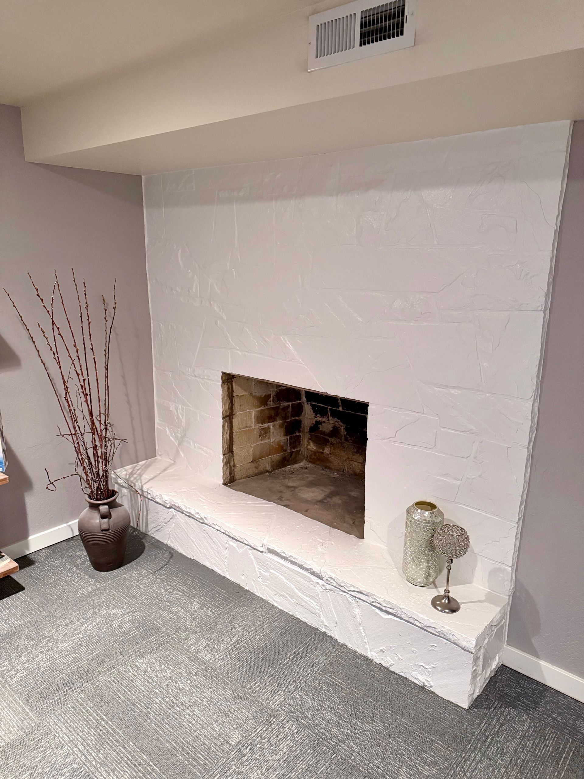 White stucco fireplace with dark interior. Light gray carpet, purple walls, decorative vase.