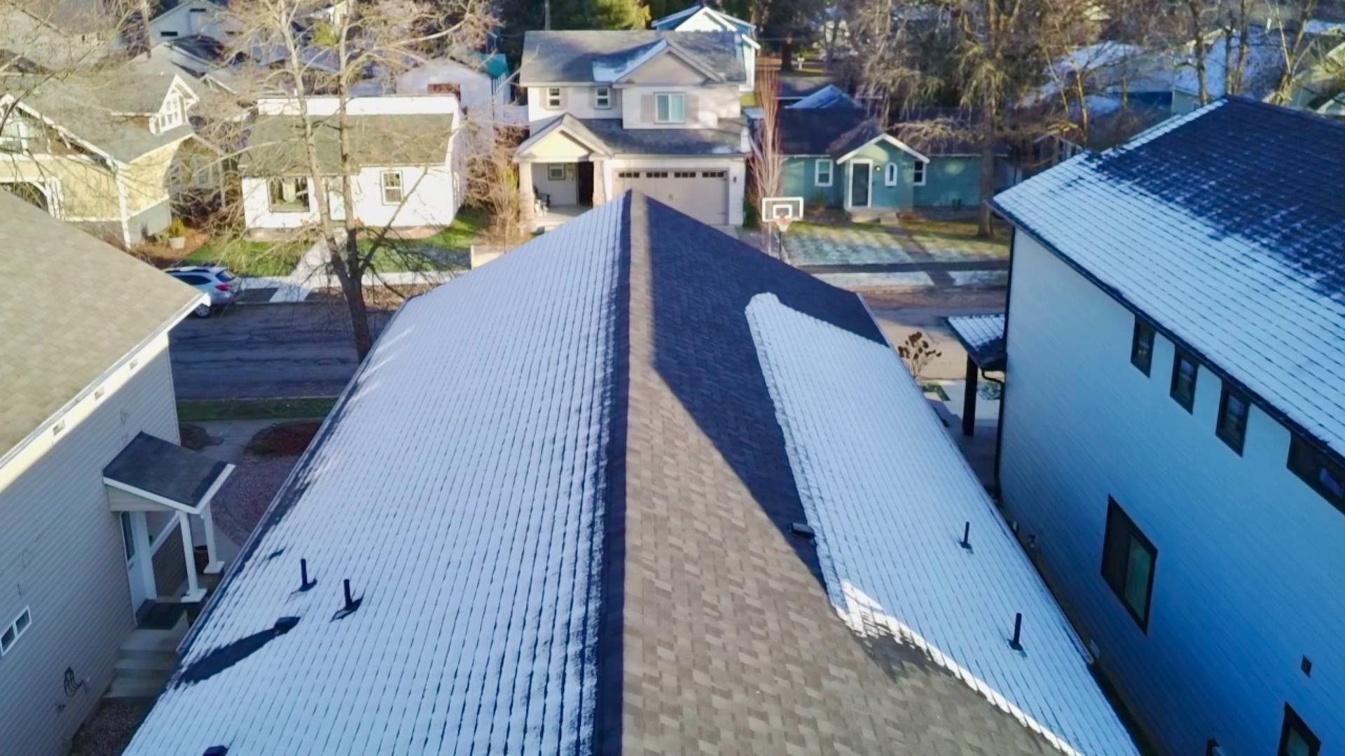 Overhead view of rooftops in a neighborhood, some covered in snow. Gray shingles, houses.