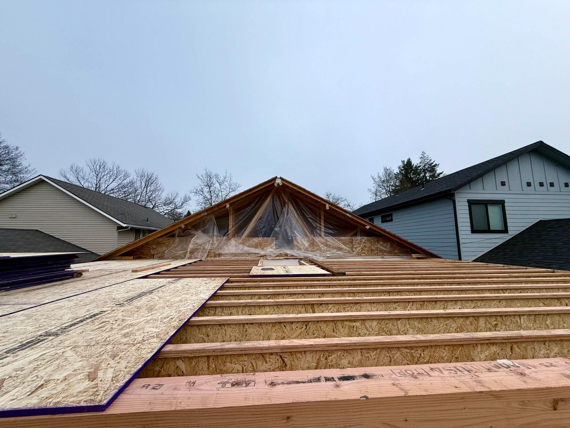 Roof under construction with exposed wooden beams, plywood, and a partial framework, with surrounding houses visible.