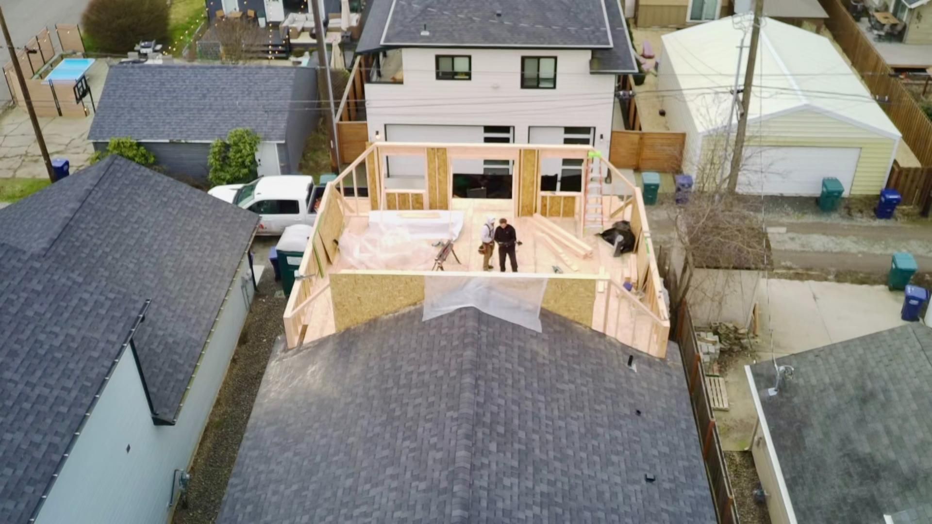 Aerial view of home construction, with workers on a partly built second story; other homes nearby.