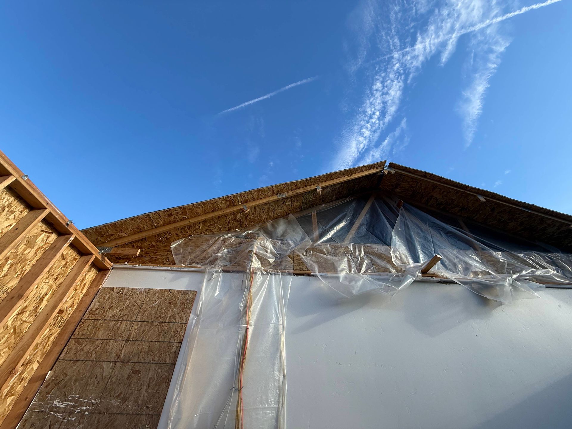 Partially constructed building with exposed wood framing, insulation, and plastic sheeting against a blue sky.