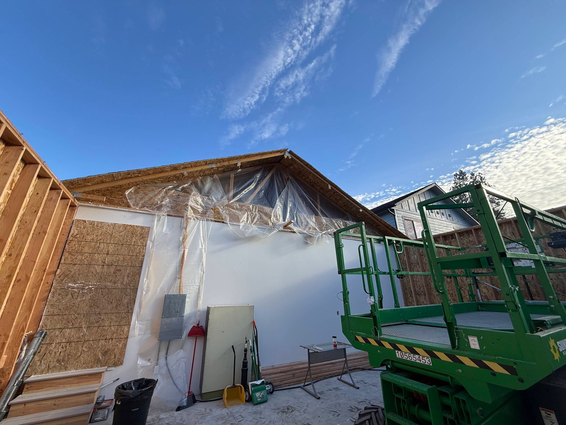 Construction site with exposed wood framing, covered in plastic, against a bright blue sky. A green lift is visible.