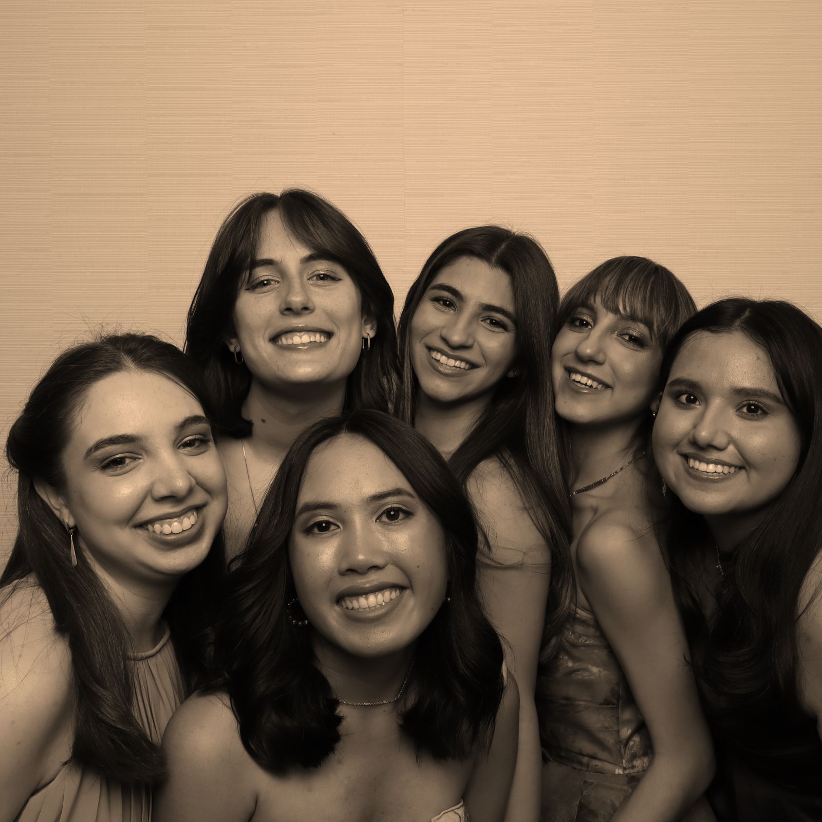 Six smiling friends pose closely together against a beige background.