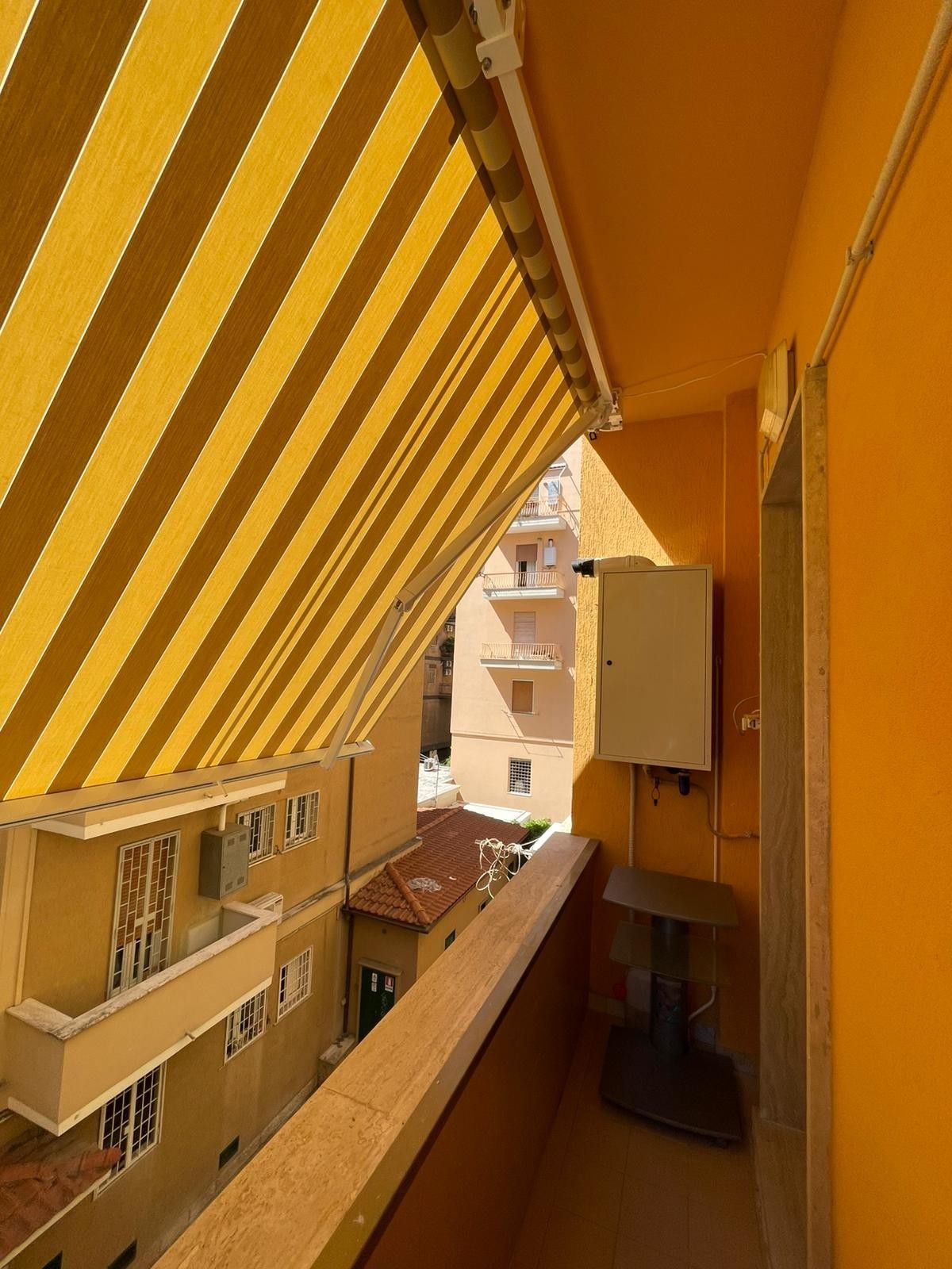 Tenda a strisce gialle su uno stretto balcone dalle pareti arancioni con vista sugli edifici.