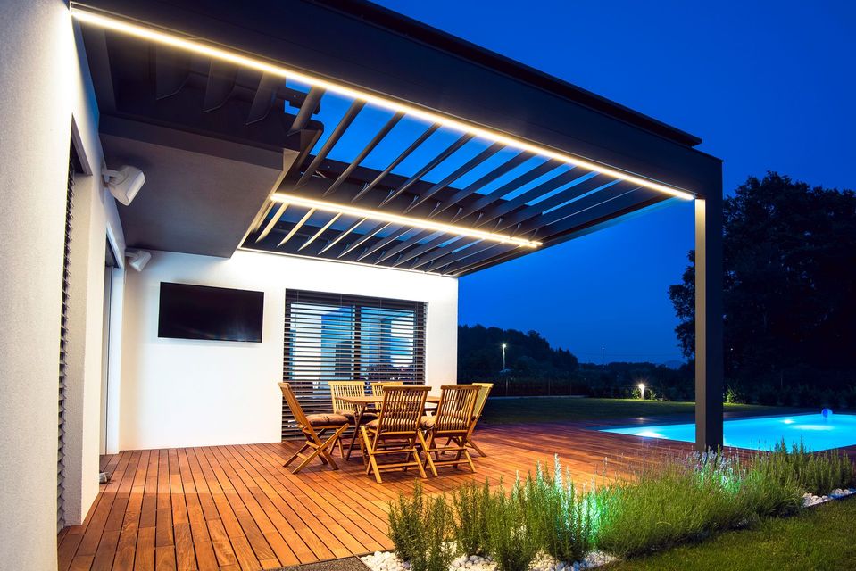 Patio esterno con pergolato illuminato, terrazza in legno, mobili e piscina.