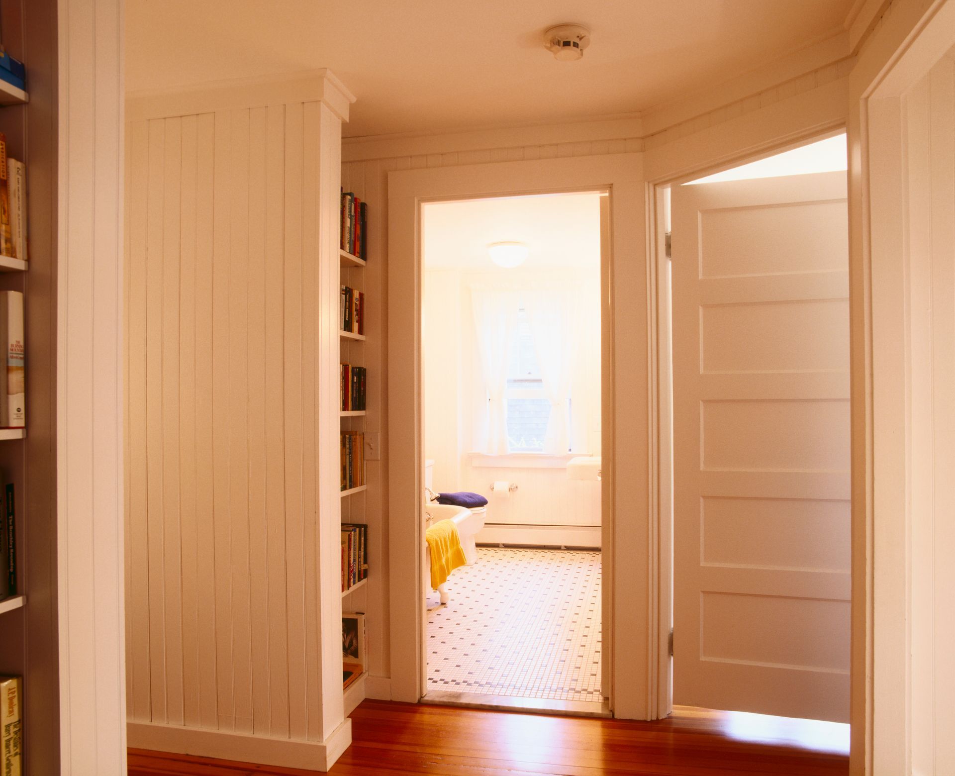 Corridoio con pareti rivestite di pannelli bianchi, libreria e porte che conducono a una camera da letto e ad altre stanze con pavimenti in legno.