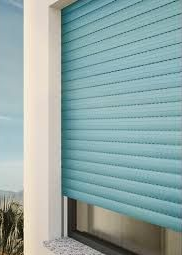 Tapparella blu con stecche orizzontali sopra una finestra. Fuori, cielo azzurro e fogliame.